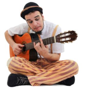 Foto de Augusto, ele está sentado no chão, tocando violão. Usa um chapeu preto pequeno, uma caçã amarelo e vermelha listrada