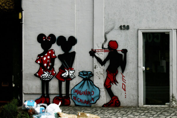 Uma parede branca exterior de um prédio, com o grafite do Mickey e Minnie de costa com pulsos amarrados, e um Saci, entre eles um saco azul escrito "Imaginário da Criança"