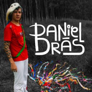 Foto de corpo de Daniel, de perfil. Ele usa uma calça branca e camiseta vermelha, em um ambiente aberto com grama e árvores ao fundo em tom cinza. Frente a ele o seu nome em branco "Daniel Brás"