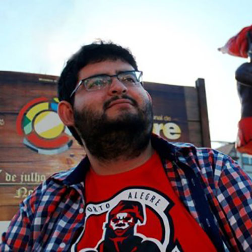 Andriolli, homem branco, com barba e cabelos cheios, óculos, em primeiro plano. Veste uma camiseta vermelha com imagem de saci e sobreposição de camiseta xadrez vermelha e azul. Ao fundo, uma estátua de saci.